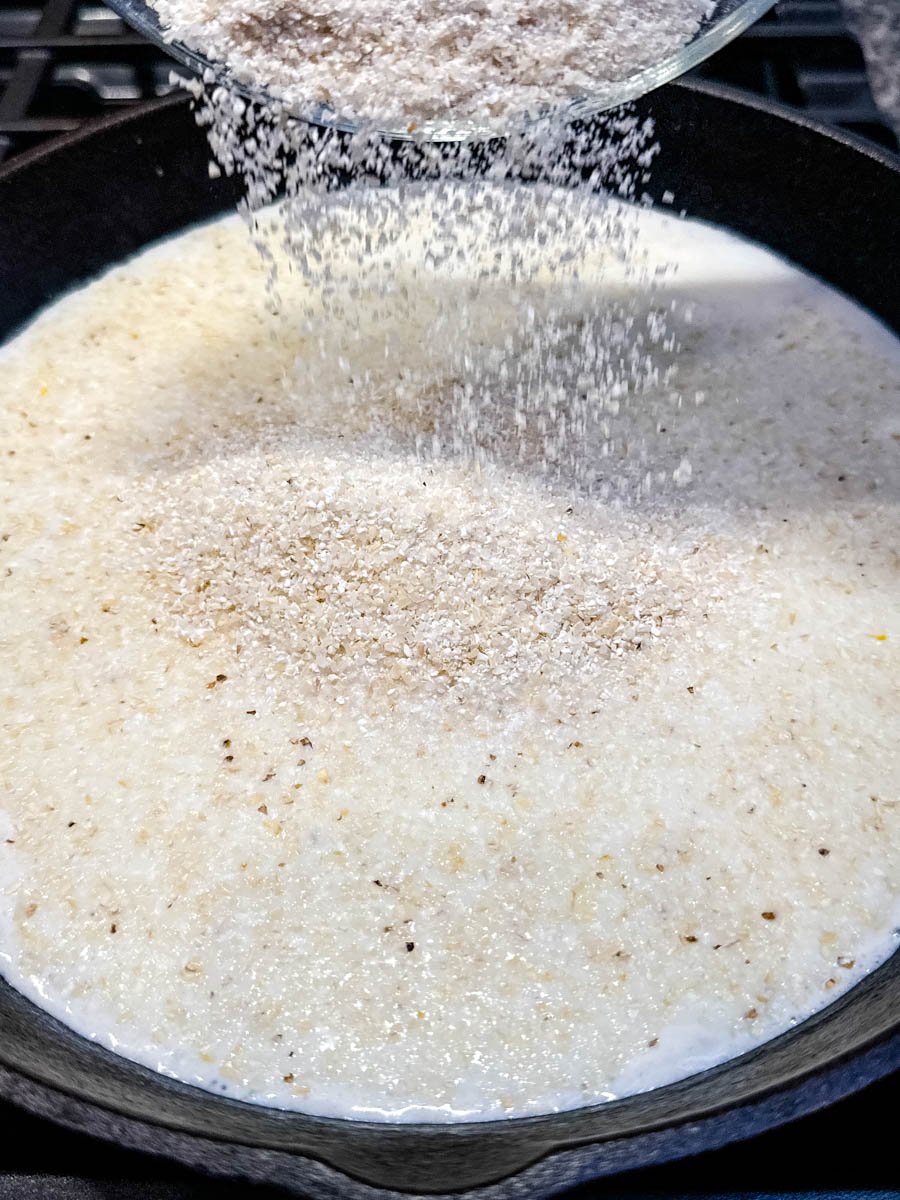 Dry stone ground grits being poured from a bowl into a cast iron skillet filled with simmering milk and water.