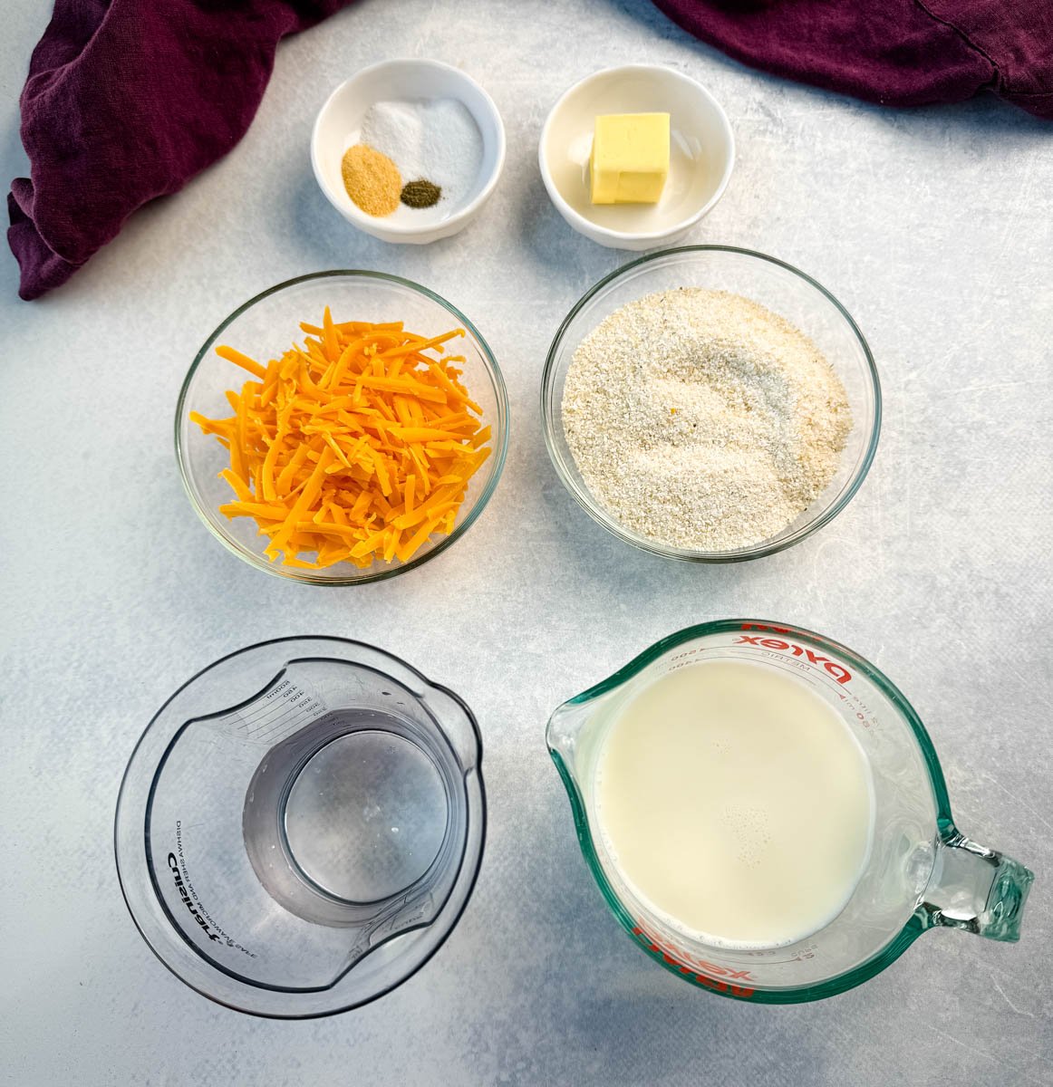 Overhead view of measured ingredients for Southern cheese grits, including shredded cheddar, dry grits, milk, butter, garlic powder, salt, and pepper.