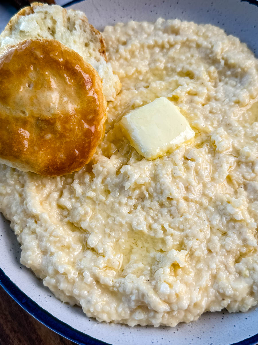 Southern-style cheese grits served in a white bowl with blue trim, finished with melted butter and a biscuit for dipping.