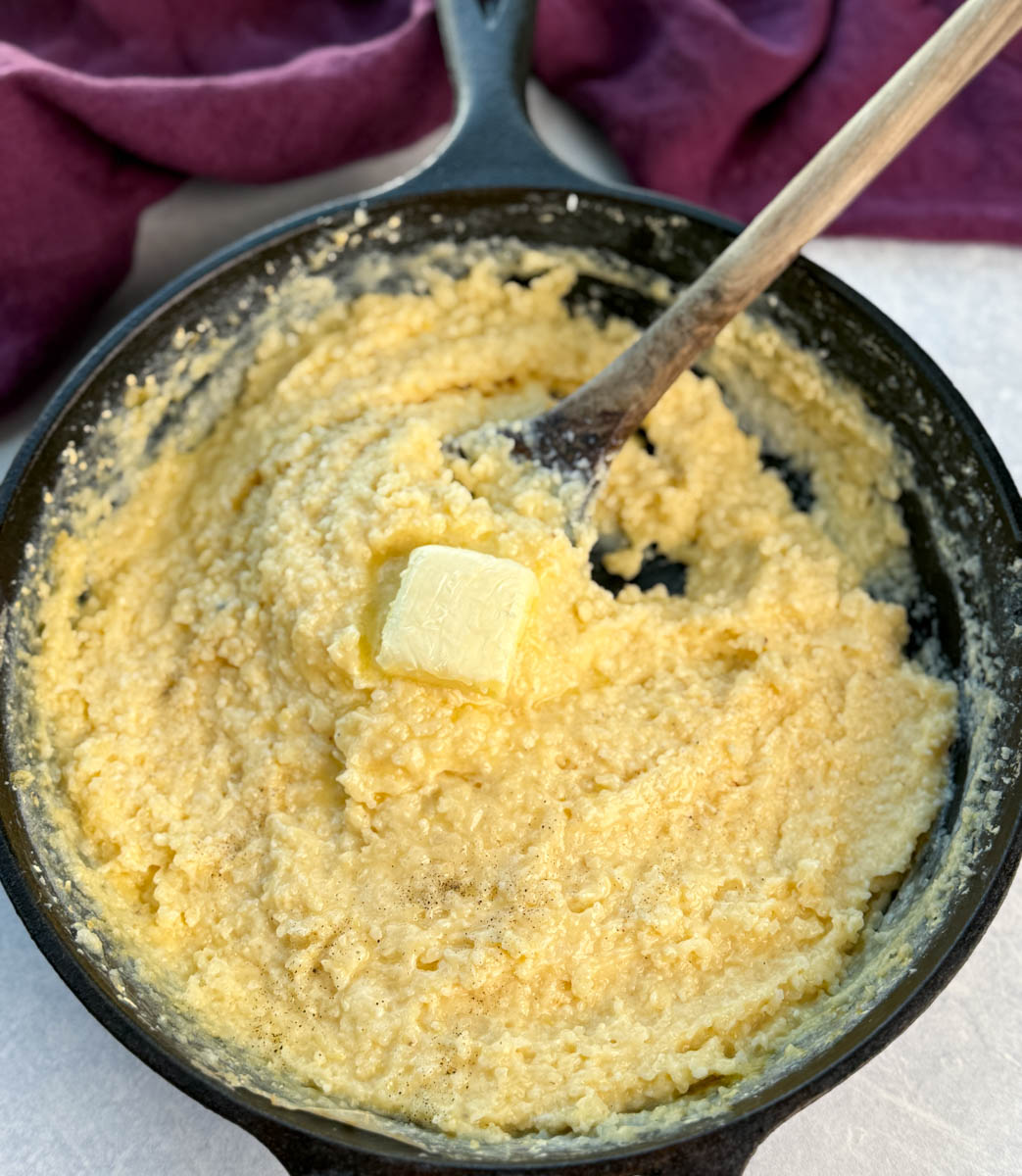 Stone ground cheese grits being stirred in a black cast iron skillet with a chunk of butter melting in the center.