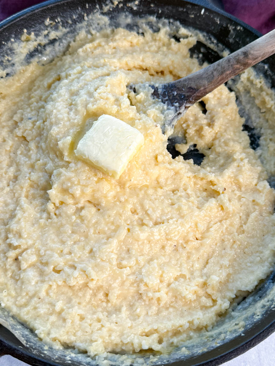 Thick and creamy cheese grits in a cast iron skillet, stirred with a wooden spoon and topped with a pat of butter.