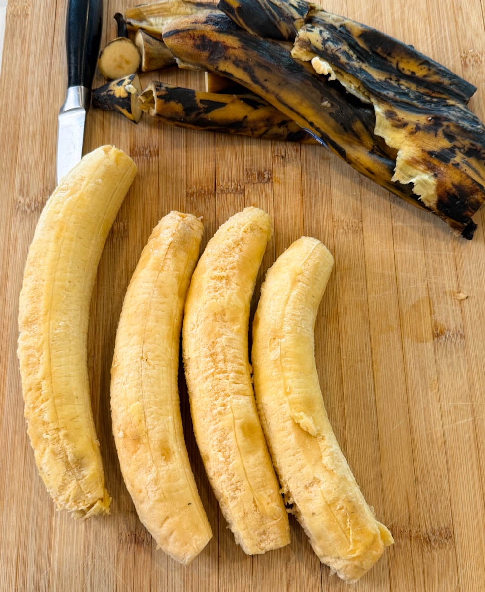 Peeled ripe plantains next to charred plantain skins on a wooden cutting board with a knife