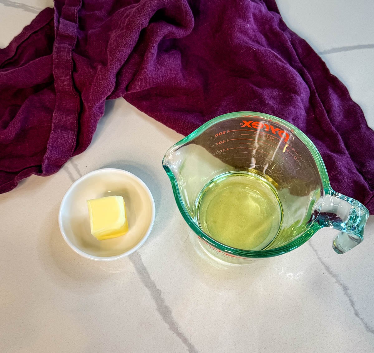 Small bowl of butter and a measuring cup of oil on a kitchen counter with a cloth in the background