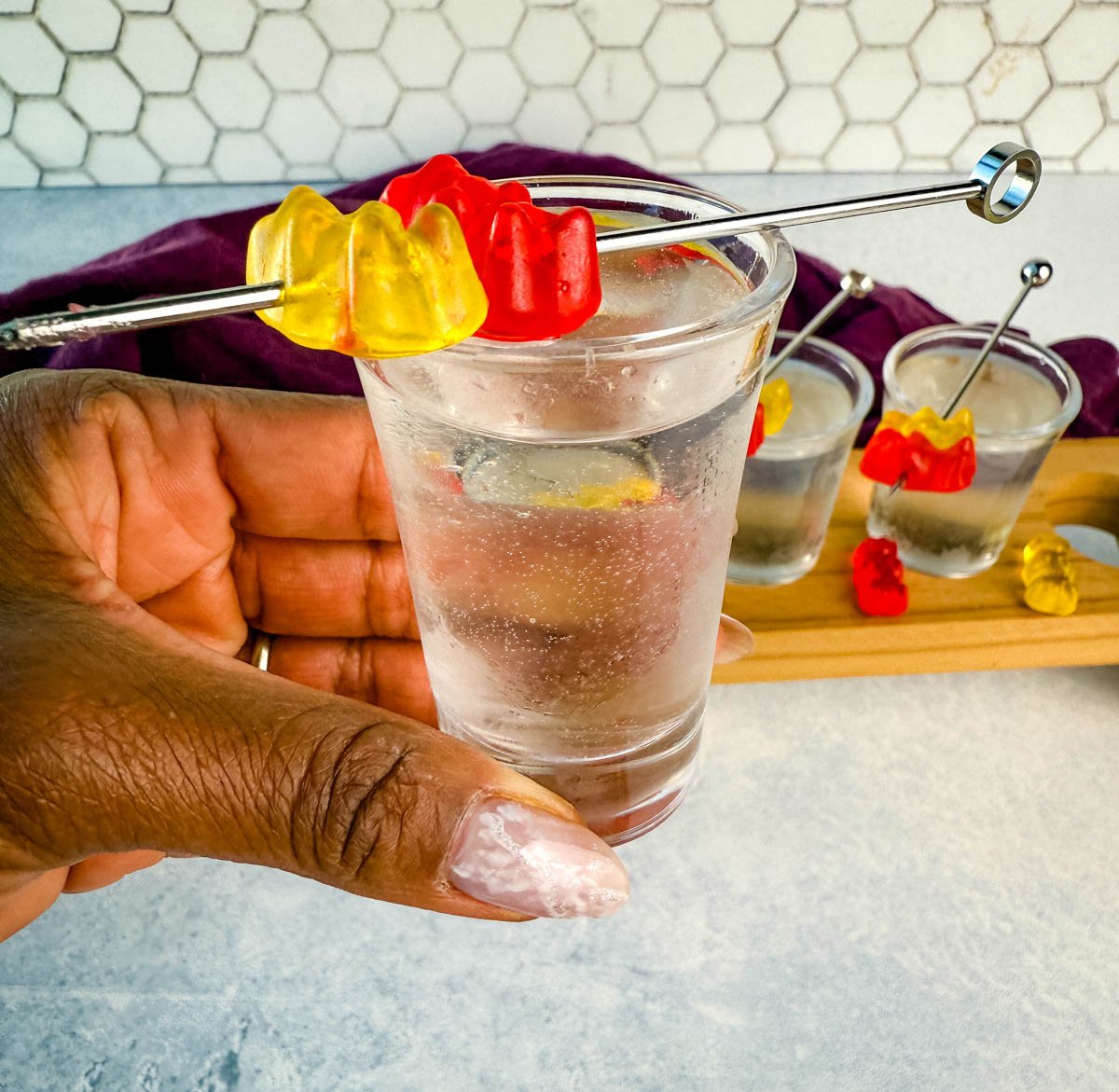 A hand holding a clear gummy bear cocktail shot garnished with red and yellow gummy bears on a metal pick. More shots and a purple napkin are visible in the background.