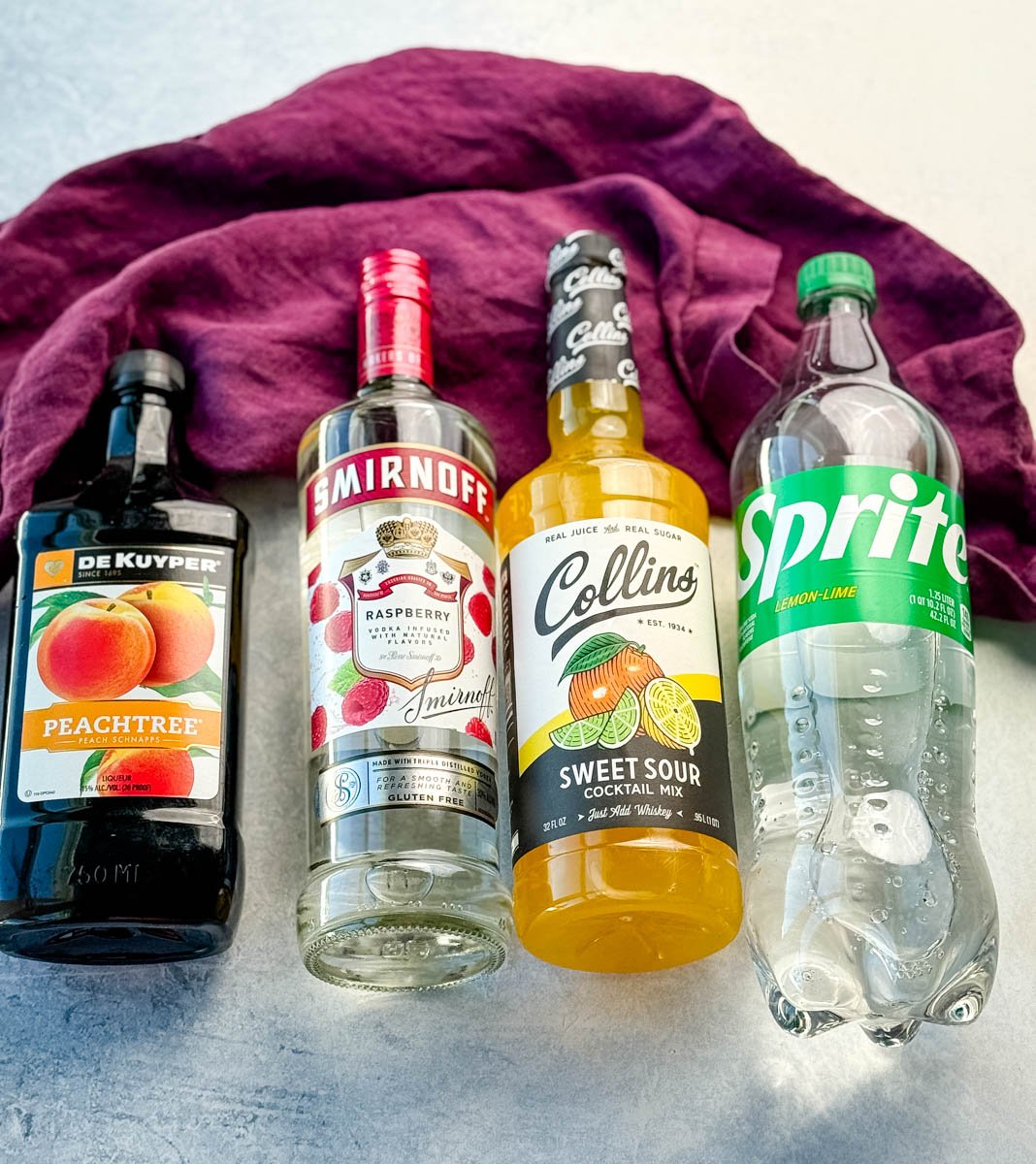 Four drink ingredients laid side by side on a light surface with a purple cloth in the background: DeKuyper Peachtree peach schnapps, Smirnoff raspberry vodka, Collins sweet and sour cocktail mix, and a bottle of Sprite lemon-lime soda.