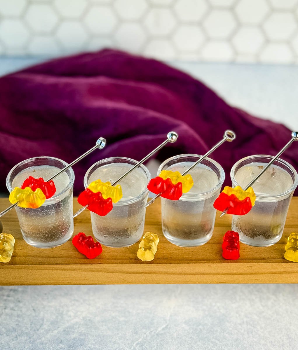 Four gummy bear cocktail shots lined up on a wooden flight board. Each shot is garnished with red and yellow gummy bears on a metal pick. A purple napkin is draped in the background.