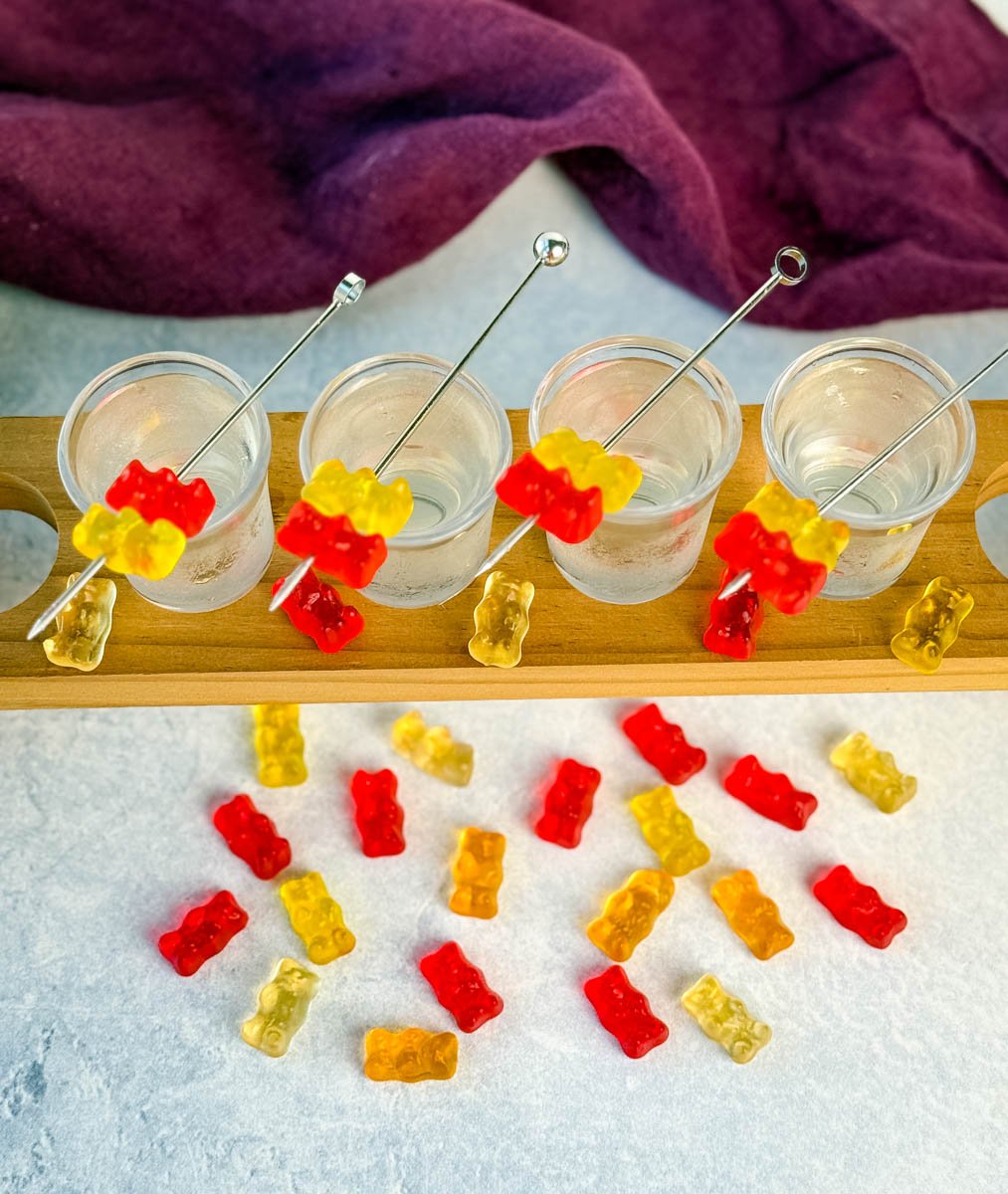 Overhead view of four gummy bear cocktail shots in a wooden flight board, surrounded by scattered red, yellow, and orange gummy bears on a light surface.