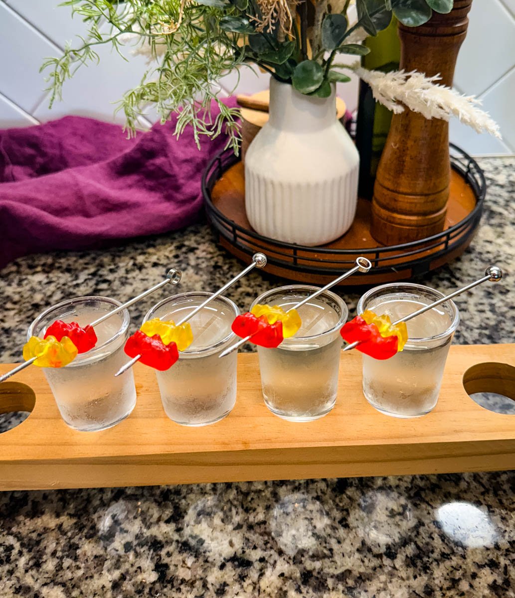 Gummy bear cocktail shots placed on a wooden flight board in a kitchen setting. Each shot is garnished with red and yellow gummy bears, and kitchen decor with greenery is visible in the background.