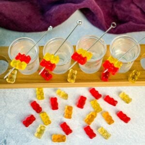 Overhead view of four gummy bear cocktail shots in a wooden flight board, surrounded by scattered red, yellow, and orange gummy bears on a light surface.