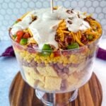 Ranch dressing being poured over the top of the layered Southern Cornbread Salad in a glass trifle dish.