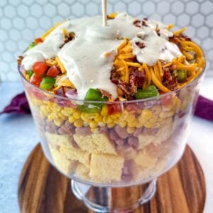 Ranch dressing being poured over the top of the layered Southern Cornbread Salad in a glass trifle dish.