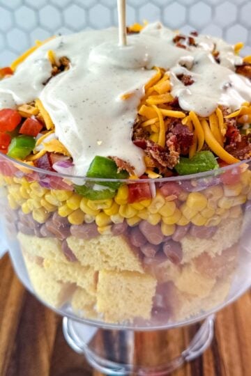 Ranch dressing being poured over the top of the layered Southern Cornbread Salad in a glass trifle dish.
