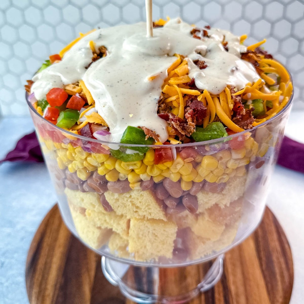 Ranch dressing being poured over the top of the layered Southern Cornbread Salad in a glass trifle dish.