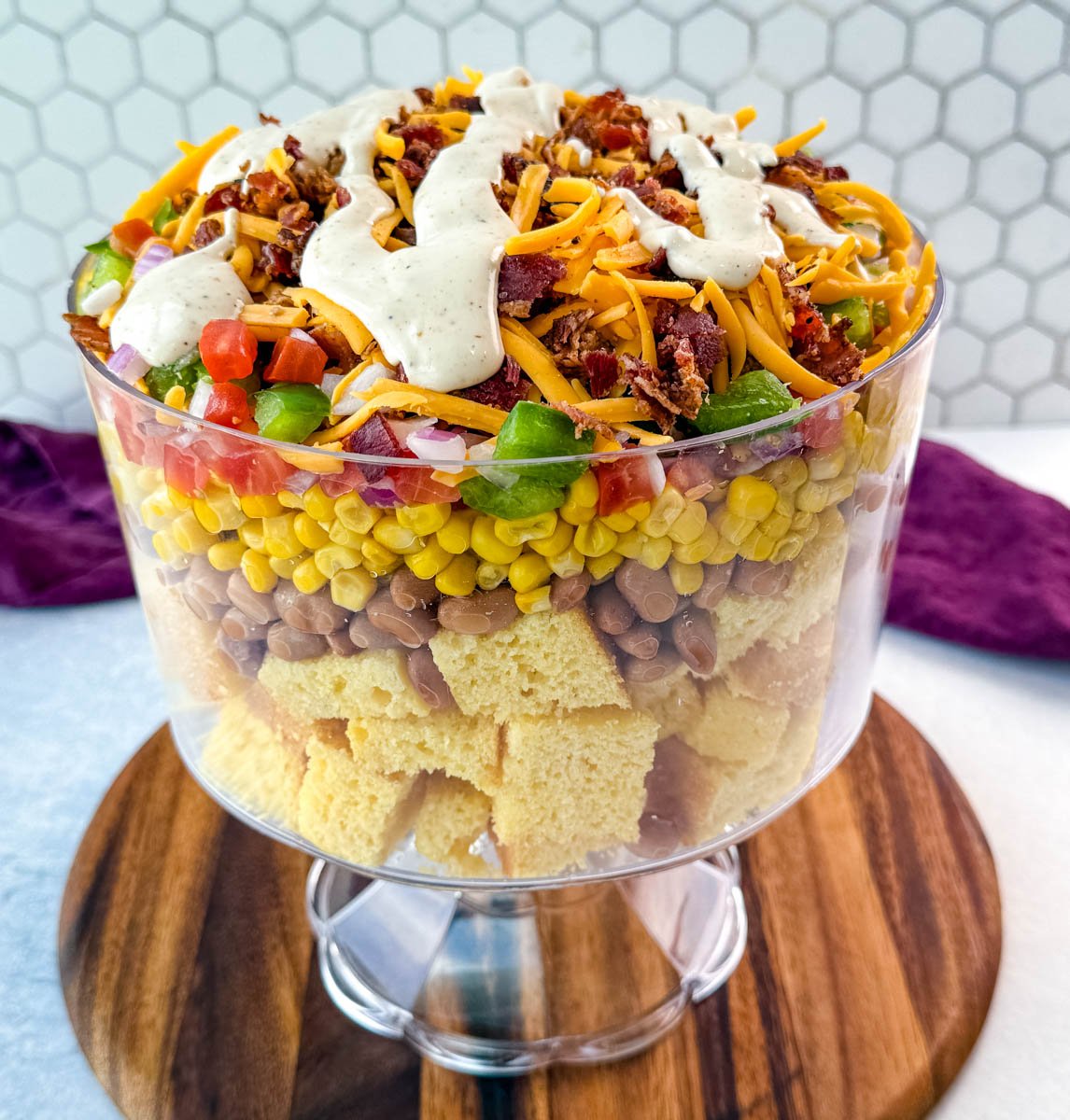 A trifle bowl layered with cubed cornbread, pinto beans, corn, diced veggies, shredded cheddar, crispy bacon, and a drizzle of ranch dressing on top.