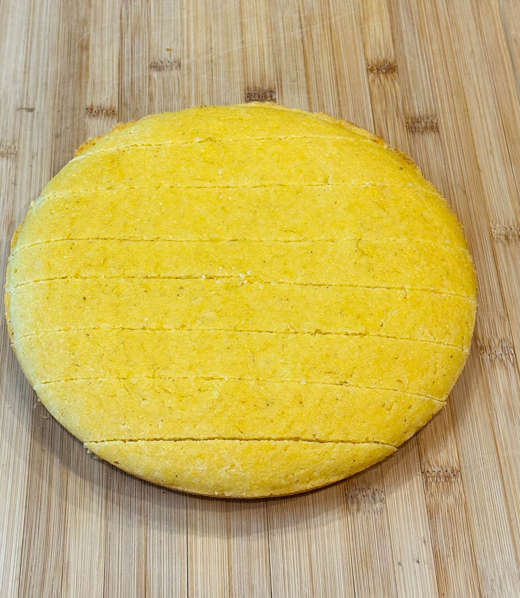 A baked cornbread round scored and ready to be cubed, sitting on a wooden cutting board.