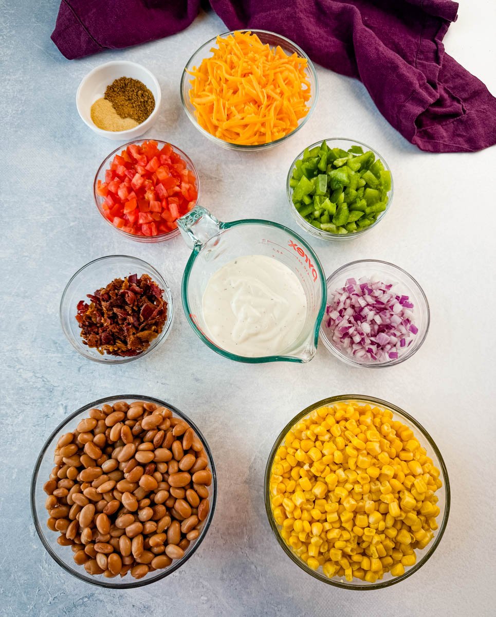 Overhead view of all the Southern Cornbread Salad ingredients laid out in bowls: pinto beans, corn, crispy bacon, shredded cheddar cheese, diced tomatoes, green bell peppers, red onion, ranch dressing, and seasoning blend with Creole seasoning and garlic powder.
