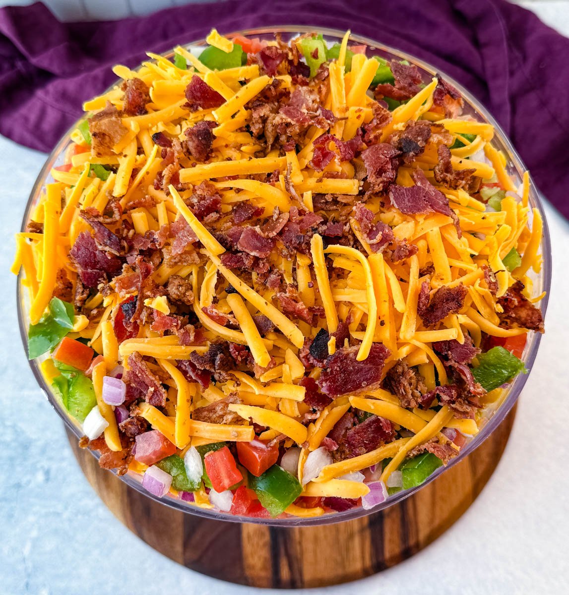 An overhead view focusing on the top layer of shredded cheddar cheese and crumbled bacon with chopped green bell peppers, tomatoes, and red onions peeking through.