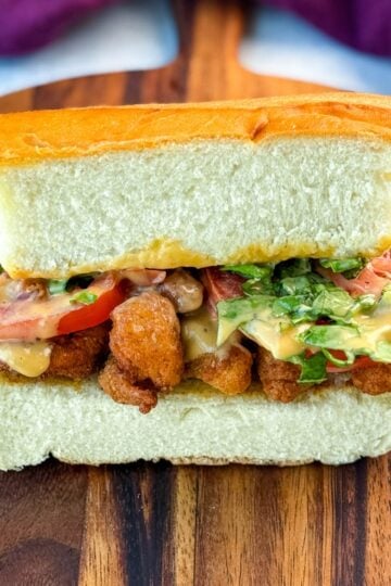 Centered image of a crispy chicken po' boy sandwich on a cutting board, showcasing layers of fried chicken, tomato, lettuce, and sauce, surrounded by chips.