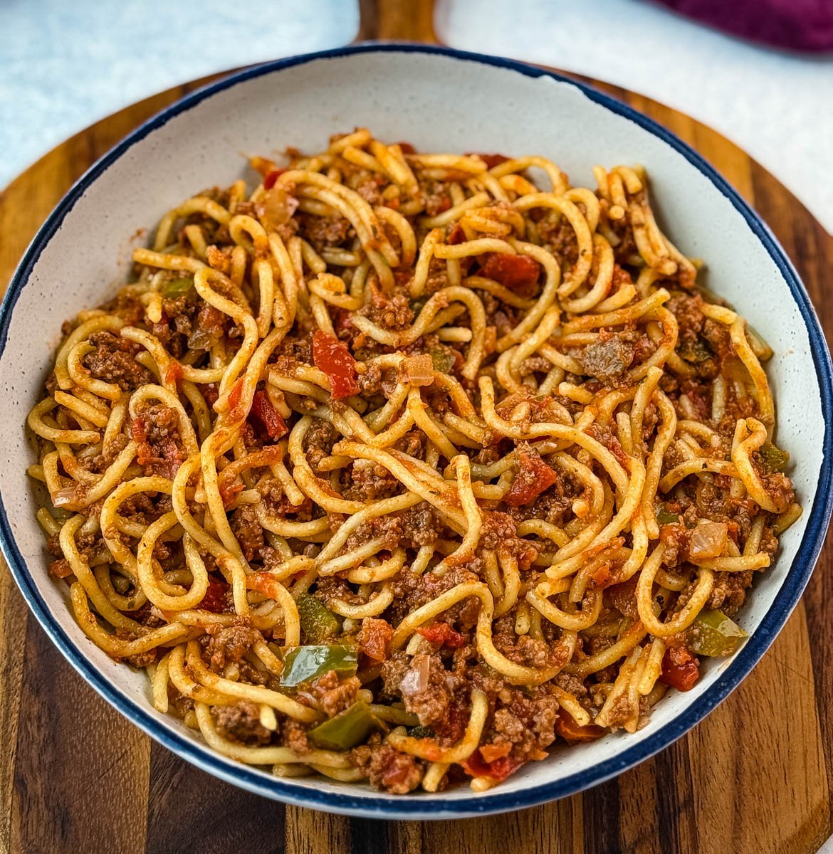 A serving bowl filled with Southern-style spaghetti, placed on a wooden cutting board, showcasing noodles mixed with ground beef, green peppers, and seasoned tomato sauce.