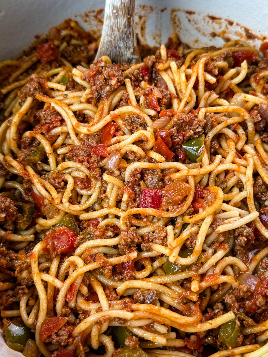 Pot of spaghetti being stirred, packed with ground beef, diced tomatoes, onions, and green bell peppers in a thick sauce.