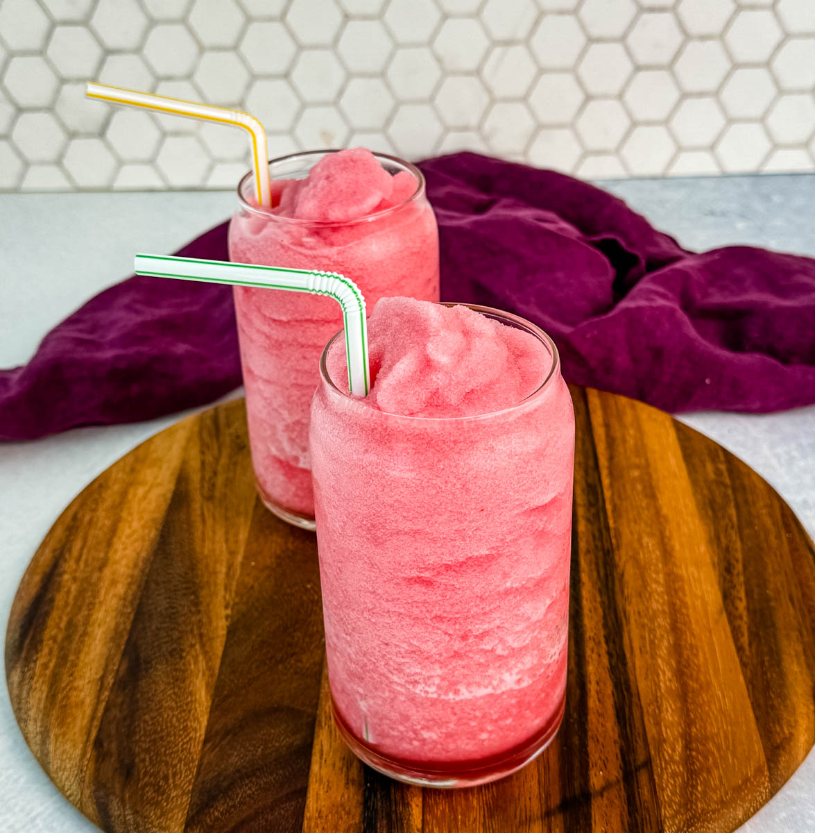 Two glasses of bright pink fruit punch slushies served with striped straws on a round wooden board, with a purple cloth in the background and a white honeycomb tile backsplash.