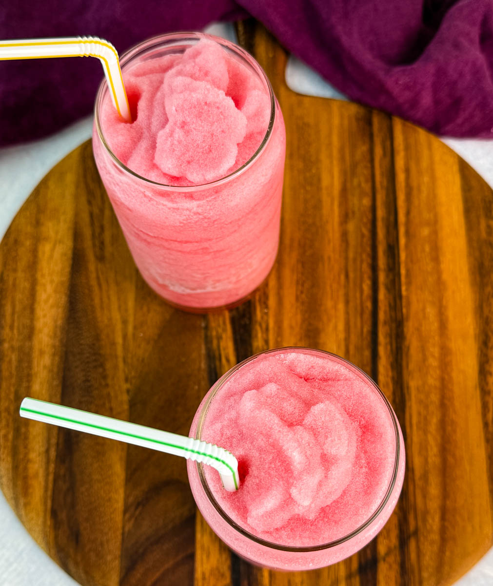 Overhead view of two glasses filled with bright pink fruit punch slush, each with a straw, sitting on a wooden tray with a purple cloth.