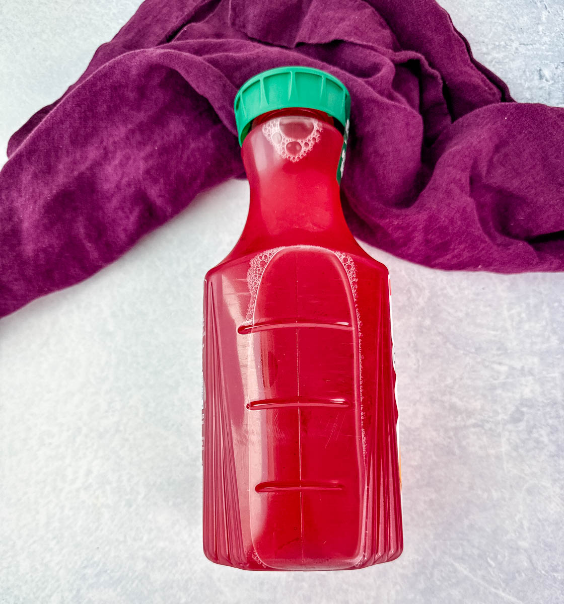A full, unopened bottle of bright red fruit punch with a green cap, placed against a light surface and a deep purple cloth backdrop.