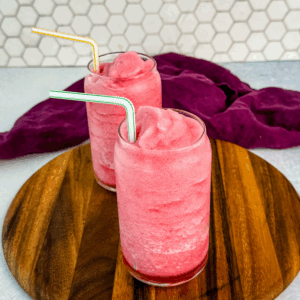 Two glasses of pink fruit punch slush placed side-by-side on a wooden tray in front of a white hexagon tile wall, each with a colorful striped straw.