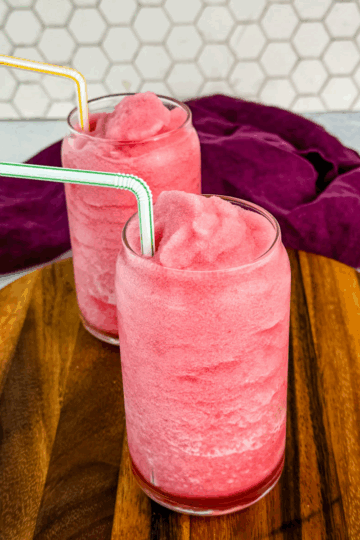 Two glasses of pink fruit punch slush placed side-by-side on a wooden tray in front of a white hexagon tile wall, each with a colorful striped straw.
