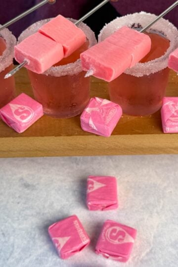 Four pink Starburst-inspired cocktail shots with sugar-rimmed glasses, each garnished with skewered pink Starburst candies. More pink Starbursts are scattered on the table in front of the drinks, with a hexagon-tiled wall and deep purple cloth in the background.