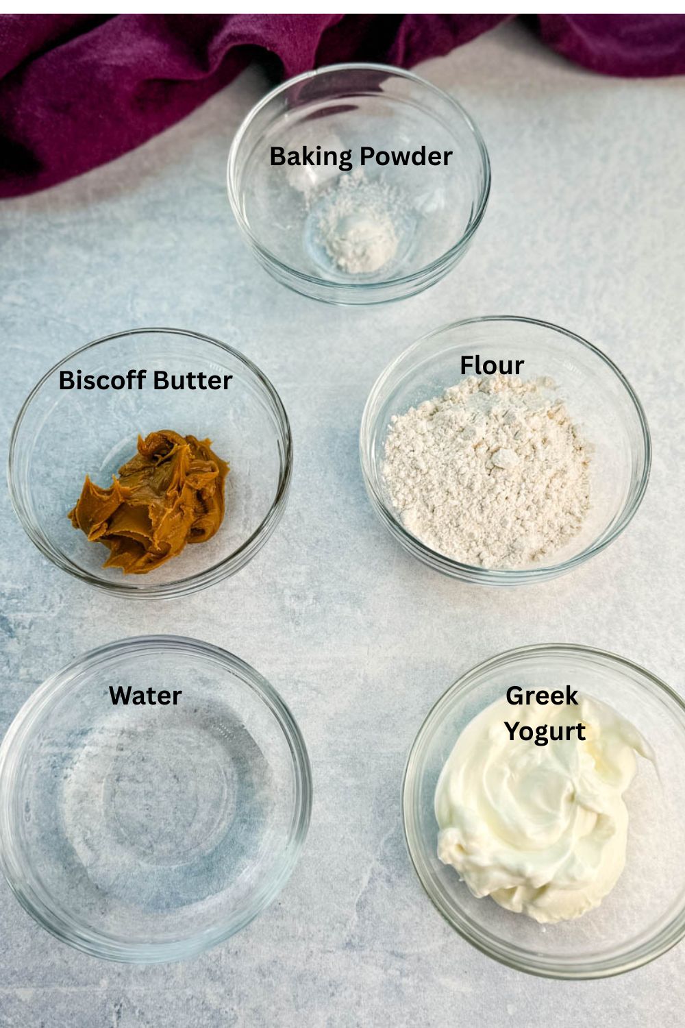 Overhead shot of glass bowls labeled with ingredients: flour, Greek yogurt, water, Biscoff butter, and baking powder.