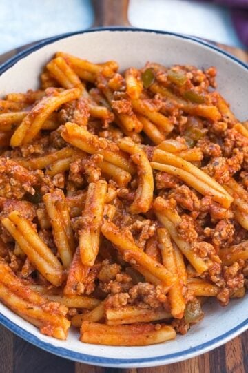 A shallow white bowl on a wooden board containing the finished pasta, with a hint of a burgundy cloth in the background, showcasing the vibrant red sauce and sausage.
