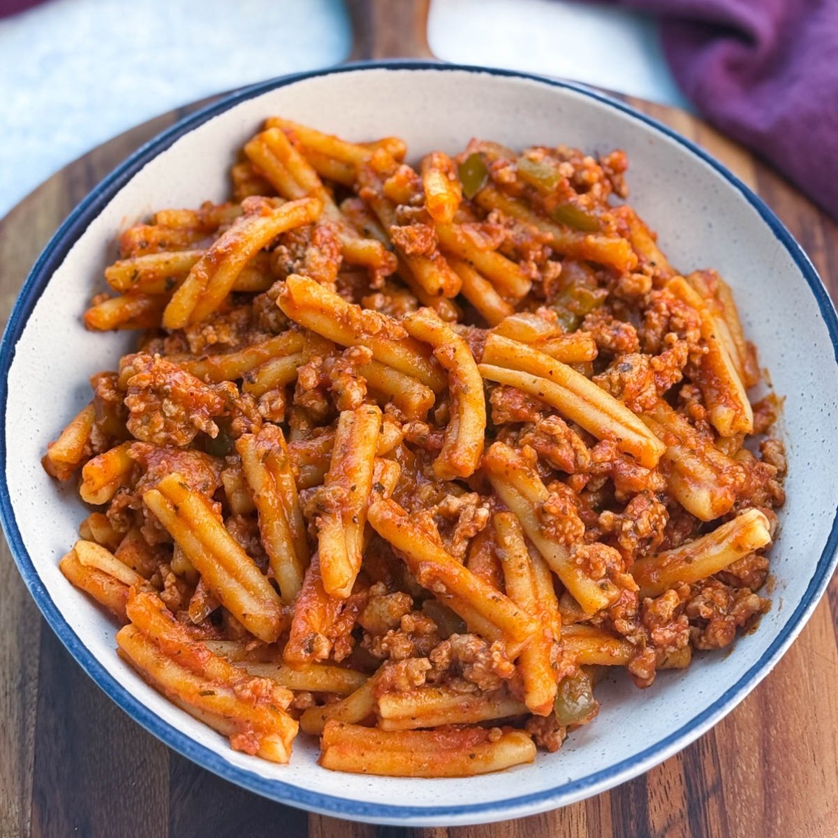 A shallow white bowl on a wooden board containing the finished pasta, with a hint of a burgundy cloth in the background, showcasing the vibrant red sauce and sausage.