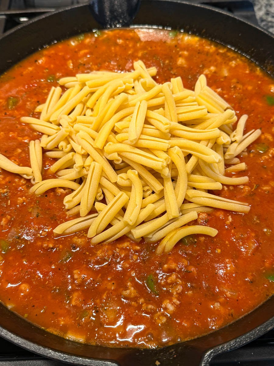 Raw casarecce noodles piled in the center of a skillet of tomato sausage sauce, just before being stirred in to cook directly in the sauce.