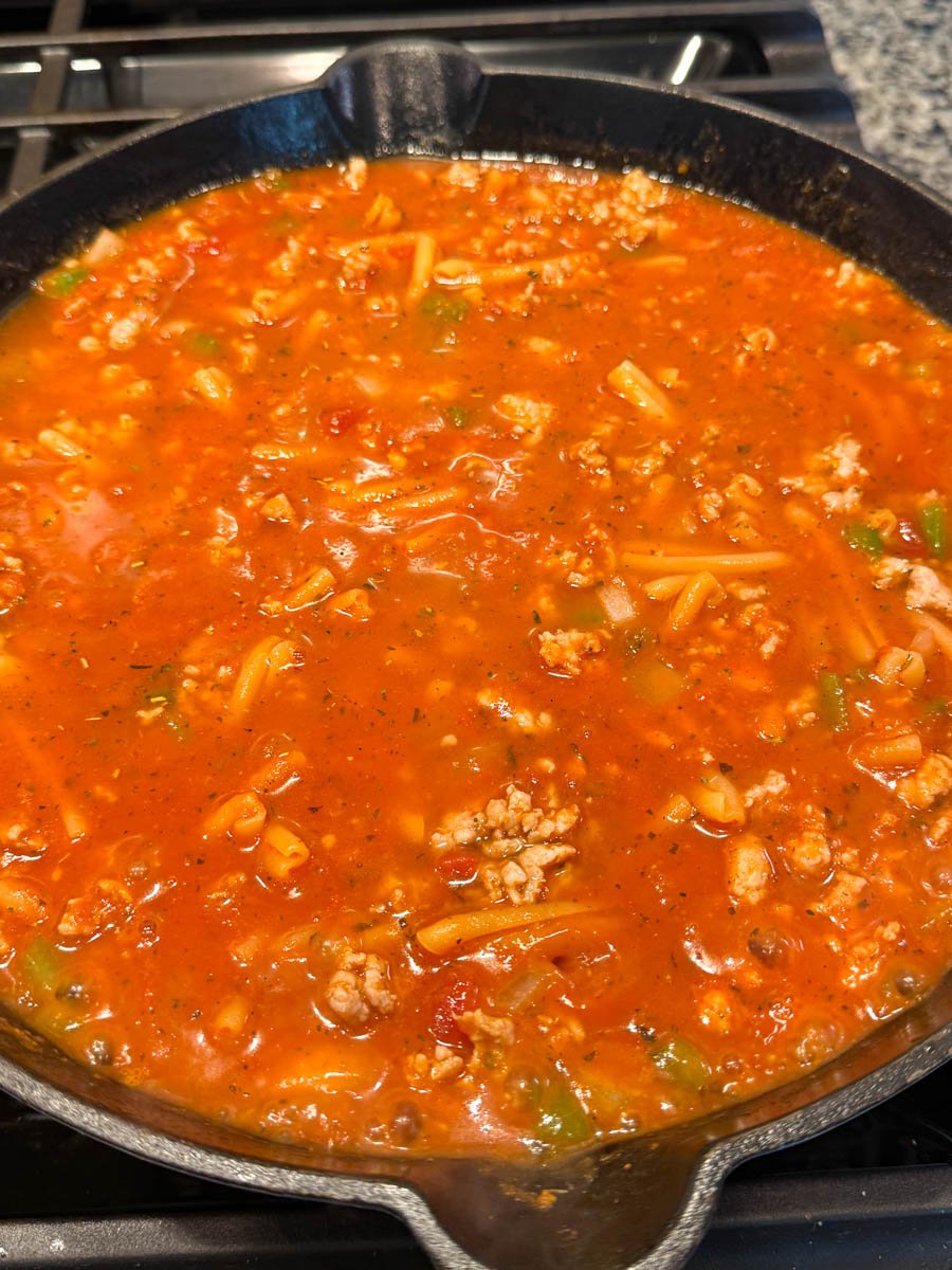 Skillet on the stovetop filled with pasta cooking in the simmering tomato-meat sauce, the noodles just beginning to soften.