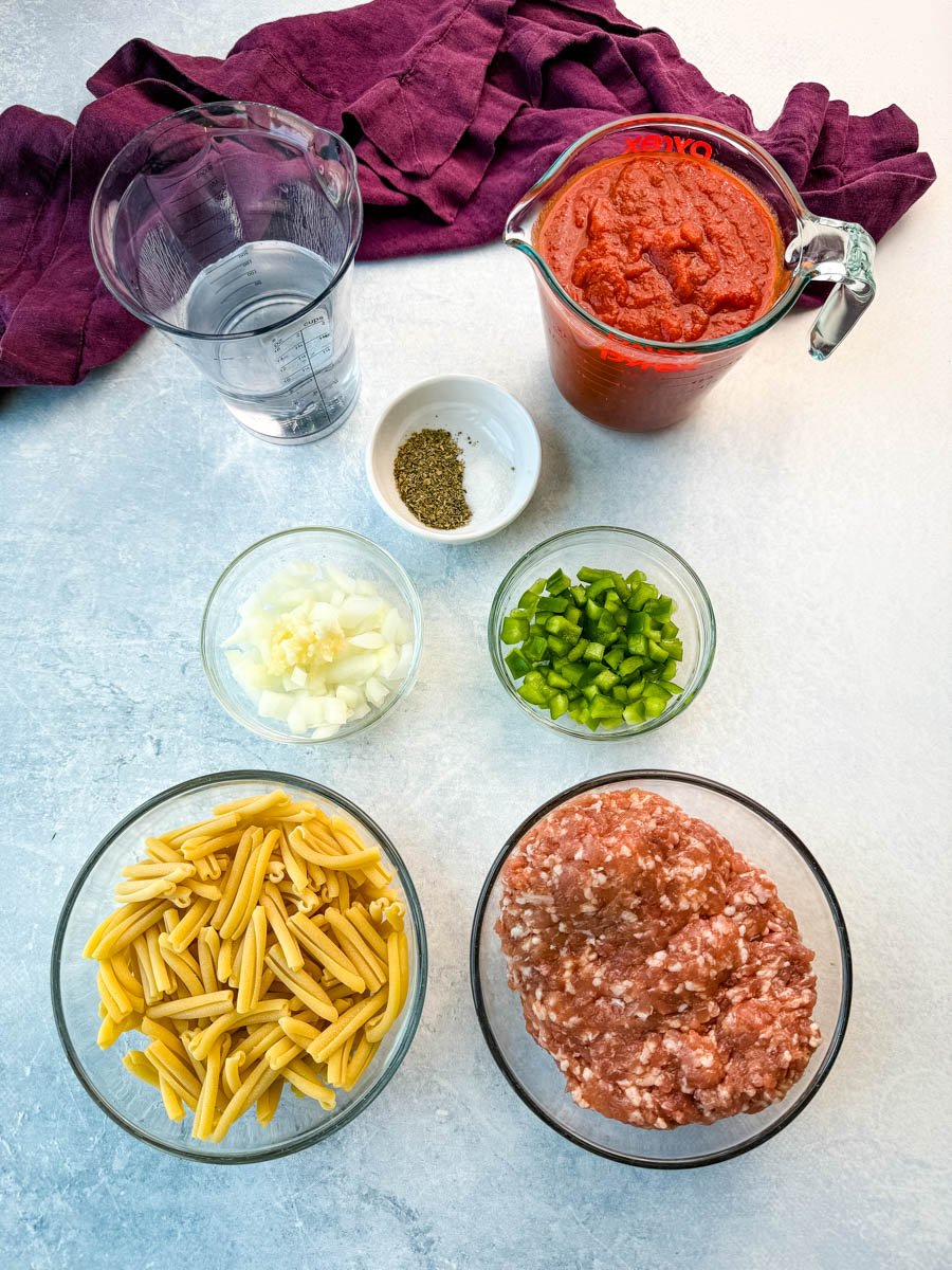 Flat-lay of ingredients on a light countertop: a clear measuring cup of water, a Pyrex pitcher filled with tomato sauce, a small bowl with dried Italian seasoning and salt, a bowl of chopped onion and garlic, a bowl of diced green bell pepper, a bowl of uncooked casarecce pasta, and a bowl of raw ground Italian sausage.