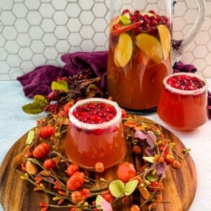 Pitcher of Thanksgiving punch with apple slices and cranberries, served with two sugared-rim glasses of punch on a round wooden board.