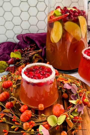 Pitcher of Thanksgiving punch with apple slices and cranberries, served with two sugared-rim glasses of punch on a round wooden board.