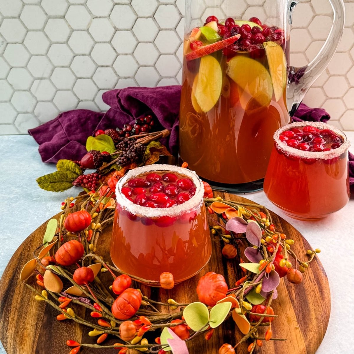 Pitcher of Thanksgiving punch with apple slices and cranberries, served with two sugared-rim glasses of punch on a round wooden board.