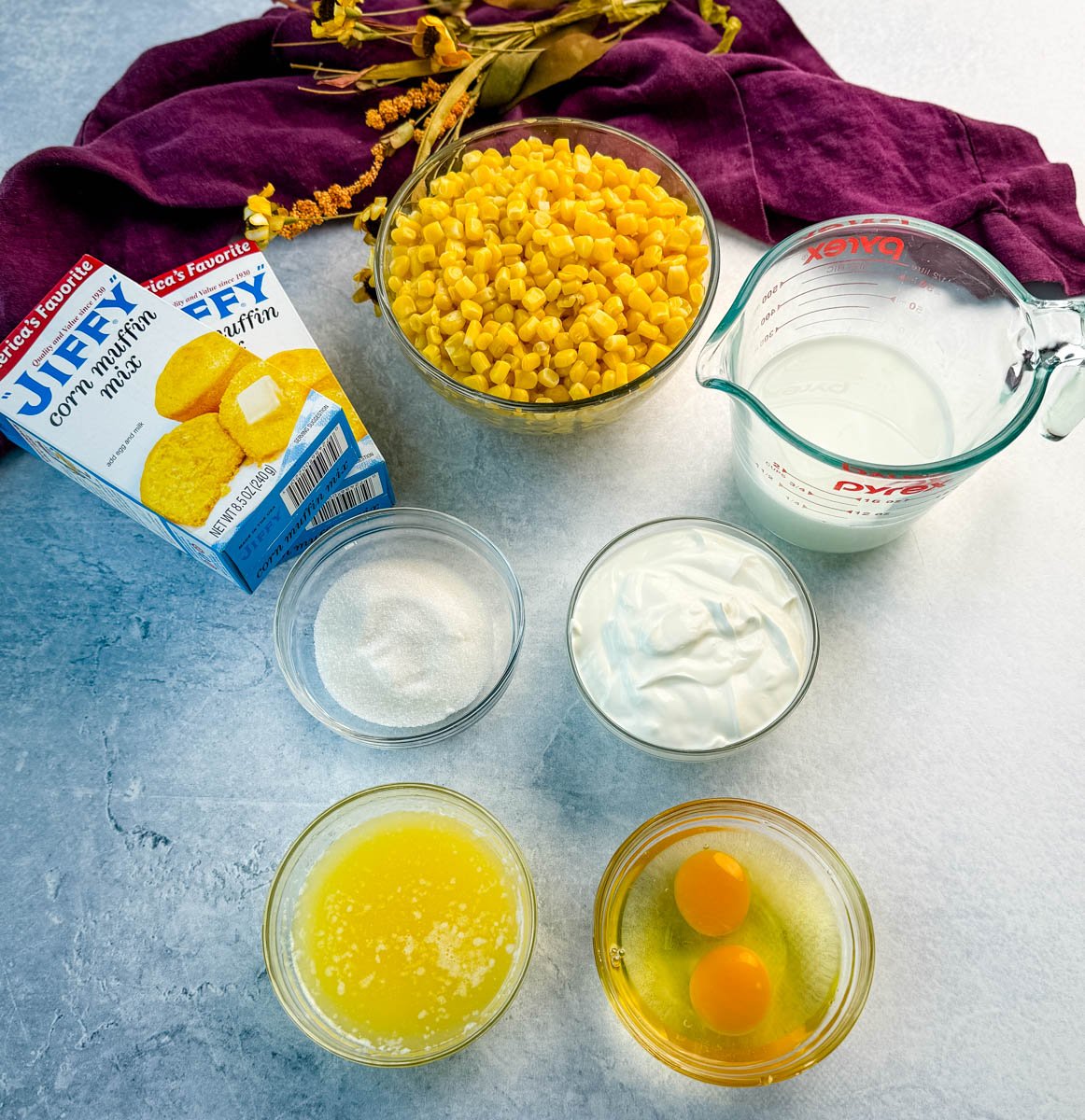 Ingredients for Jiffy corn casserole arranged on a light gray surface, including Jiffy corn muffin mix boxes, a bowl of corn kernels, sugar, sour cream, melted butter, eggs, and milk, with a purple cloth in the background.
