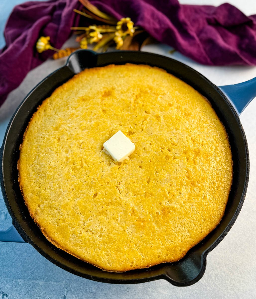 Whole baked Jiffy corn casserole in a black cast iron skillet, topped with a pat of butter in the center, purple cloth and flowers in the background
