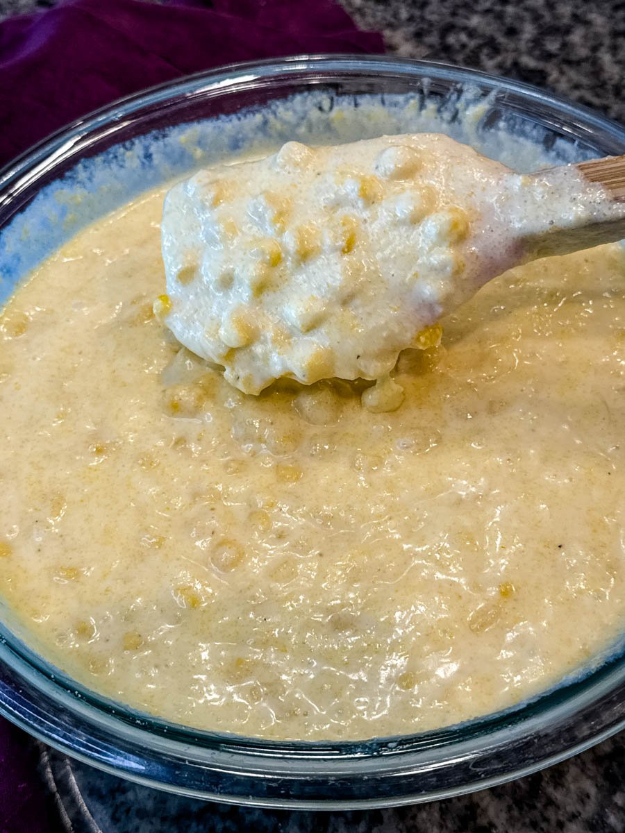 Glass mixing bowl filled with Jiffy corn casserole batter, corn kernels visible, with a wooden spoon lifting a portion.