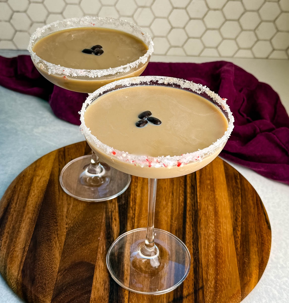Two coupe glasses filled with creamy peppermint espresso martinis, each rimmed with crushed peppermint and garnished with three coffee beans, displayed on a wooden board with a deep purple cloth in the background.