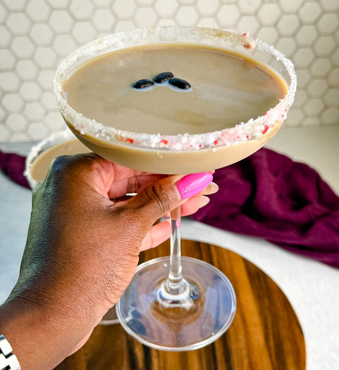 Hand holding a coupe glass with a finished peppermint espresso martini, rimmed with crushed peppermint and garnished with coffee beans, with another glass blurred in the background.