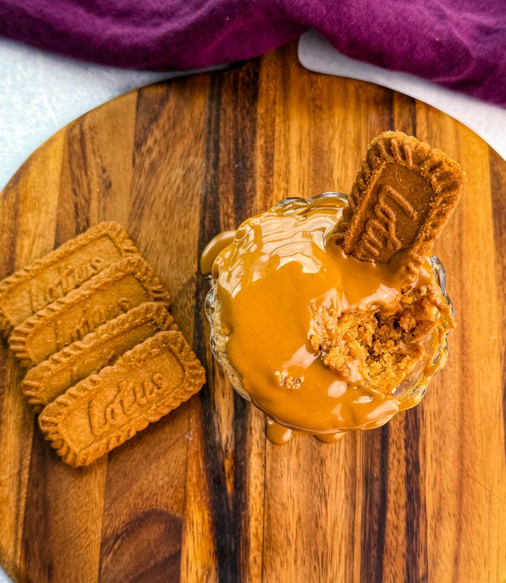 Glass dish filled with Biscoff protein mug cake, topped with melted Biscoff spread and garnished with a whole Lotus cookie, with extra cookies on the side.