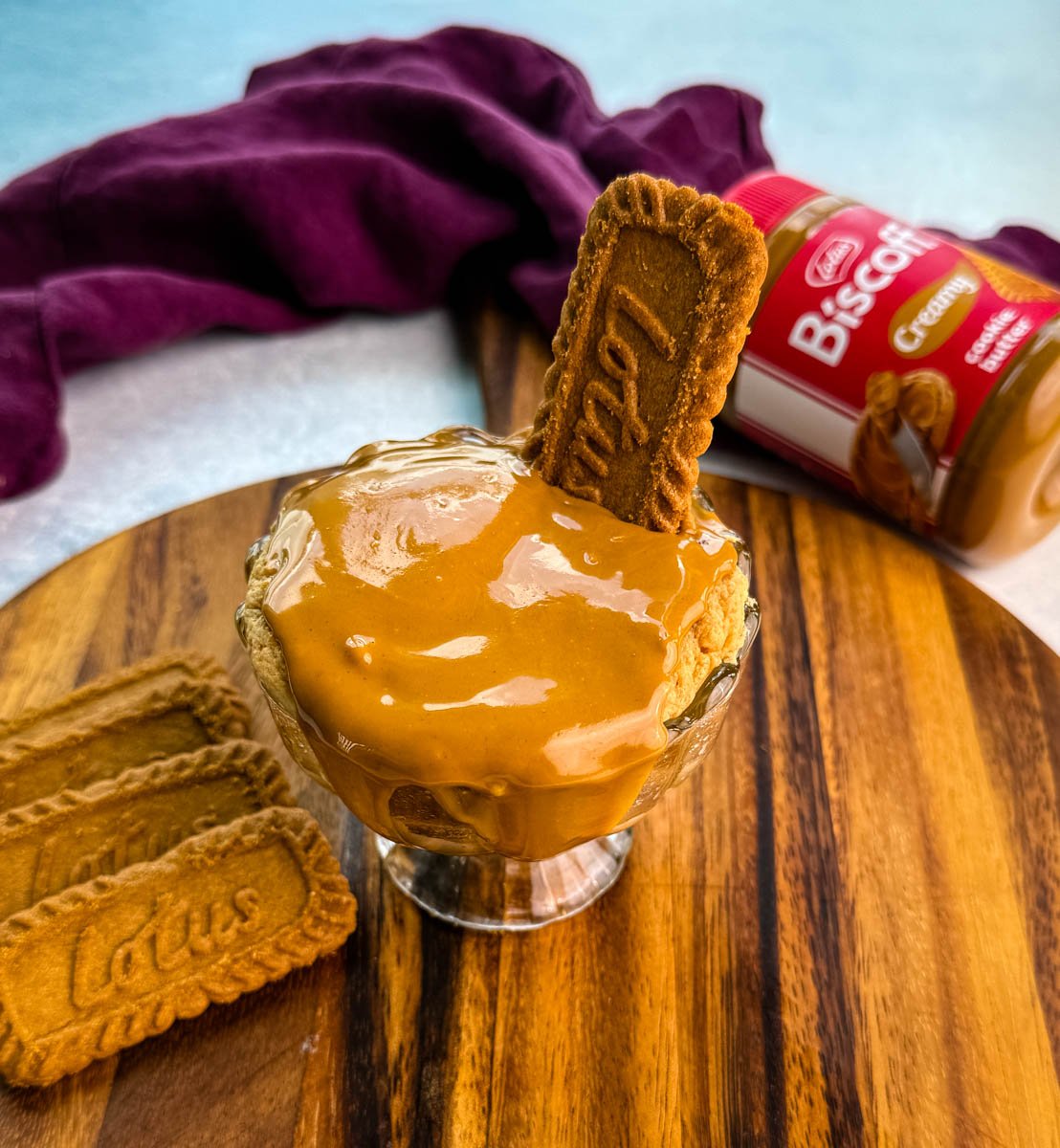 Glass dish of Biscoff protein mug cake topped with melted cookie butter and a Lotus cookie, placed beside a Biscoff spread jar and extra cookies.