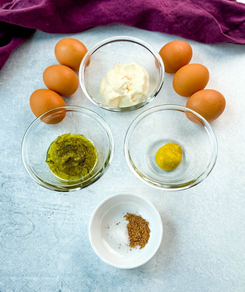 Ingredients for deviled eggs laid out on a light surface: mayonnaise, relish, mustard, seasoning, and brown eggs.