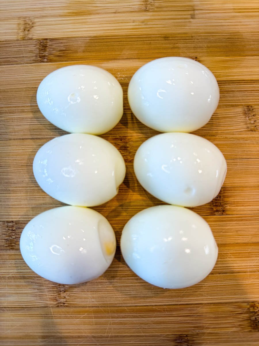 Six peeled hard-boiled eggs on a wooden cutting board.