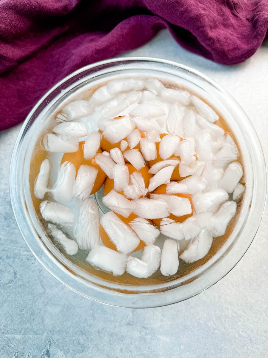 Brown eggs in a glass bowl filled with ice water after boiling, with a purple cloth in the background.