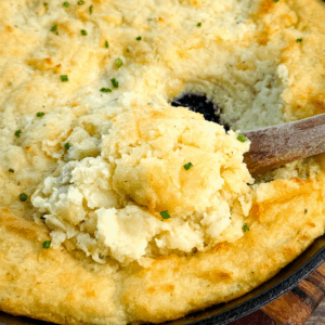 Close-up of a cast-iron skillet filled with golden, fluffy baked mashed potatoes topped with chives, with a wooden spoon scooping out a serving.